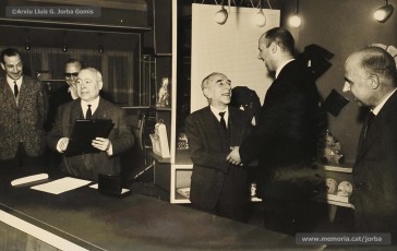 (12/14) Any 1965. Homenatge a Lluis Torra Pujol (1900-1968) amb motiu de la seva jubilació. Aquest empleat havia treballat 50 anys a Can Jorba, a l’administració de la secció de queviures. L’acompanyen Pere Jorba Puigsubirà, a la seva dreta, i Lluís G. Jorba Gomis, a la seva esquerra. Pere Jorba Puigsubirà féu el parlament en nom de l’empresa.