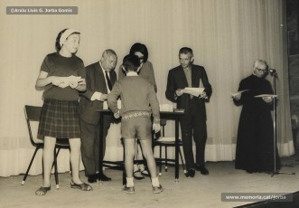(1/19) Octubre de 1966. Acte de fi de curs 1965-66 de les sessions de cinema infantil. Lliurament del record d’assistència per Pere Jorba Puigsubirà, president del Consell d’Administració de Magatzems Jorba, Dolors Ponsà, empleada de Cal Jorba, i el pare Ferran Torra Sisquella SJ (1893-1990), director de les Congregacions Marianes.