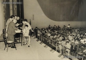 (9/19) Octubre de 1966. Acte de fi de curs 1965-66 de les sessions de cinema infantil. Lliurament del record d’assistència per Pere Jorba Puigsubirà, president del Consell d’Administració de Magatzems Jorba, Dolors Ponsà, empleada de Cal Jorba, i el pare Ferran Torra Sisquella SJ (1893-1990), director de les Congregacions Marianes.