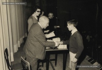 (14/19) Octubre de 1966. Acte de fi de curs 1965-66 de les sessions de cinema infantil. Lliurament del record d’assistència per Pere Jorba Puigsubirà, president del Consell d’Administració de Magatzems Jorba, Dolors Ponsà, empleada de Cal Jorba, i el pare Ferran Torra Sisquella SJ (1893-1990), director de les Congregacions Marianes.