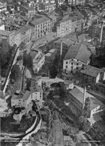 Vista aèria del torrent de Sant Ignasi i del salt dels Gossos, obtinguda pel fotògraf manresà Josep Gaspar i Serra des d'un aeroplà l'any 1926. A l'esquerra, de dalt a baix, les fàbriques de l'Areny, de les Fontetes i del Salt. En la part superior esquerra de la fotografia, el carrer de Codinella amb la casa Asols. Aquest torrent havia estat utilitzat des de l'inici del procés industrialitzador com a indret preferent de localització fabril. Però el seu aprofitament hidràulic (les aigües provenien de les escorrialles de la sèquia) s'havia de combinar amb l'ús de la màquina de vapor, ja que, sobretot a l'estiu, baixava pràcticament sec. El torrent, a més, era un focus permanent d'infecció perquè, tal com ens ho recorda Josep Oliveras, s'anà convertint en el col·lector d'una part de la xarxa del clavegueram de la ciutat. Així, de manera insistent, apareixien a la premsa escrits de denúncia sobre el mal estat en què es trobava el torrent, especialment en el seu tram final, al baixador de Sant Marc. Al novembre del 1895, el Diario de Avisos ressenyava que «todo aquello es un verdadero charco; y ni saltando acá y acullá, y donde largos rodeos, se logra apenas poder atravesar aquel pantano. Esto parece imposible y sin embargo es tal como decimos. Si se deja permanecer en aquel estado tan concurrido sitio, qué van a decir los forasteros de nuestra ciudad?». En aquest sentit, la preocupació per la salubritat pública, pròpia dels higienistes del tombant de segle, també la tingué l'alcalde Maurici Fius i Palà, qui ordenà el cobriment del torrent des de la placeta de Sant Ignasi fins a la font de l'Obac. Les obres, tanmateix, es quedaren aturades i no van ser acabades fins l'any 1935.
Procedència: Revista "Ciutat" (Arxiu Comarcal del Bages)