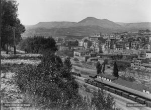 Perspectiva de la ciutat presa des del turó de Santa Caterina. Un paisatge sec contrasta amb el casc urbà que es perfila a l'altra banda del riu, on es delimiten amb nitidesa un tram del passeig del Riu, els molls de l'estació del Nord, i la fàbrica de cal Cots. Més enllà, sobresurt l'edifici de les sederies Balcells - actual seu del Conservatori Municipal de Música- amb la seva característica torre quadrangular i els darreres de les cases dels carrers d'Arbonés i del Llop; al fons, el Collbaix, sorprenentment nu de vegetació al tombant de segle XX i l'antiga fàbrica dels Panyos. 
Procedència: Col·lecció Ramon Cornet i Arboix. (Fotografia de Lluís Cornet i Vivar)