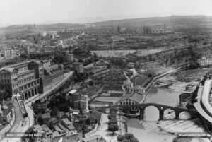 El sector del Pont Vell tal com es podia admirar des del campanar de la Seu, als anys vint del segle XX. En aquesta àmplia panoràmica s'observen, a mà dreta, la carretera d'Esparreguera; a l'altra banda del riu, la fàbrica del Pont Vell, dedicada als filats i teixits de cotó i regentada en aquests moments per la companyia Argemí, Gallifa i Vila. Aquesta fàbrica, adossada al Pont Vell, fou bastida entre el 1859 i el 1862 sobre l'antic emplaçament d'un molí fariner, el Molí Nou. A l'esquerra, l'edifici de la Cova i la Casa d'Exercicis. Al peu, les cases del carrer i de la capella de Sant Marc, i al darrere, l'inici del camí de la Cova. L'últim edifici perceptible és el convent de Santa Clara. Seguint a l'horitzó, l'ampla balconada de Sant Pau, seu en l'actualitat d'un conjunt residencial i, més enllà, sobre la carena de cal Gravat, d'un polígon industrial. I pel dessota, es perceben les obres del desmunt de la via dels Ferrocarrils Catalans, que a partir del 1926 comunicarien la ciutat amb Barcelona.
Procedència: Arxiu Jaume Pons i Agulló