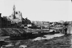 Panoràmica parcial de Manresa des del peu de la riba dreta del riu, just a sota del pont de Sant Francesc, al principi dels anys vint del segle XX, amb la façana de ponent de la Seu a mig reformar i l'agulla piramidal, l'imposant edifici de la Cova de Sant Ignasi i el convent de Santa Clara. 
Procedència: Arxiu Jaume Pons i Agulló