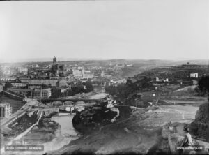 La ciutat vista des de la carretera de Can Massana, dita també del Bruc, a l'indret conegut amb el nom de Coll-Manresa. Datada entre els anys vuitanta i el tombant del segle XX. De manera preeminent sobresurten la basílica de la Seu i la Cova de Sant Ignasi. A la dreta, la torre de Santa Caterina. Al peu del riu Cardener, en la confluència amb la muralla de sant Francesc, la fàbrica del pont de fusta. A la riba dreta del Cardener, el característic pont fumat, per sobre de la via del ferrocarril que comunica Lleida amb Manresa.
Procedència: Arxiu Comarcal del Bages