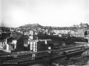 Vista parcial de la ciutat feta a principi de segle XX des de la riba esquerra del riu Cardener, baixant pel camí del Congost. En primer terme, observem les diverses vies del ferrocarril econòmic de Manresa-Berga. Al centre, els tallers de cal Canyís, ubicats a la confluència entre el passeig del Riu i el passatge d'en Coma. En aquest mateix indret, anys més tard, el fabricant Fermí Roca i Coma hi construiria la fàbrica de filats i teixits amb les seves dependències, la torre de l'amo, habitatges unifamiliars per a obrers, etc. Al fons, el castell de Puigterrà. Al peu del turó del castell, es veu l'església parroquial de Sant Pere Màrtir i, al fons, a la dreta, l'església del Carme i la caserna, ampliada al tombant de segle XX.
Procedència: Arxiu Comarcal del Bages