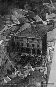 Fotografia aèria de la Plaça Major i de la Casa de la Ciutat l'any 1926. Al fons, l'antic Jutjat, la porta lateral de la Seu, la casa del Paborde i la Rectoria. La plaça ofereix un aspecte molt concorregut: les parades del mercat, gent que manté una actitud expectant davant els treballs d'ornamentació de la façana de l'edifici de l'Ajuntament. Aquest era i és el punt neuràlgic de la ciutat: plaça del mercat de l'horta manresana, punt de manifestacions i protestes i de celebracions polítiques, l'indret preferit per escoltar les bandes de música dels regiments destacats a la caserna del Carme; lloc, en darrer terme, on es rebien amb tots els honors les personalitats que visitaven la ciutat. En aquests moments, hom considerava que fer el mercat en aquesta plaça pública era un signe d'endarreriment, i s'afirmava que «tot el progrés que podem mostrar al nostres visitants en l'ordre de la construcció, comerç i fabricació, i fins, si voleu, de mesures i reformes urbanes en determinats indrets de la ciutat, resta eclipsat quan es topa amb aquest relleu de la venda al carrer, i ens col·loca en una situació d'inferioritat manifesta, total, davant de les ciutats que posseeixen un Mercat tancat a propòsit per aquest servei públic».
Procedència: Revista "Ciutat" (Arxiu Comarcal del Bages)