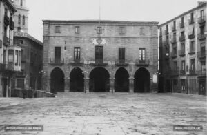 Perspectiva de la Plaça Major i la Casa de la Ciutat. Al llindar del balcó principal, una placa recorda que aquesta és la "Plaza de la Constitución". Aquest majestuós edifici fou bastit entre els anys 1739 i 1777 sota les ordres del mestre d'obres Jeroni Torrents, per reemplaçar la casa del Consell de la Ciutat, que des del 1658 ocupava el mateix solar i que l'any 1713 havia estat incendiat. Durant molt de temps, a la planta principal es trobava el despatx de l'Alcaldia, les oficines i el Saló de Plens (inaugurat el 1885); i, a la segona planta, l'Arxiu, la Biblioteca i el Museu Municipal, serveis creats l'any 1896 sota l'alcaldia de Francesc Gallifa i Gomis. Avui dia, tot l'edifici és destinat a dependències municipals.
Procedència: Arxiu Llorenç Gamisans i Villaplana (fotografies de Llorenç Gamisans i Miquel Ausió).