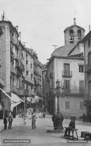 Detall de la Plana de l'Om, amb el campanar de l'església de Sant Miquel al fons, i en primer terme la sastreria Sellarès, on fins el 1995 van funcionar els Magatzems Sant Miquel.
Procedència: Arxiu Jaume Pons i Agulló.