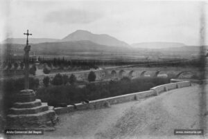 El Pont Nou i la creu que porta el seu nom en una imatge de principi del segle XX. A l'altra banda del riu, el cementiri i la muntanya del Collbaix. Antigament, aquesta era una creu de pedra, però un temporal esdevingut l'any 1765 l'esmicolà en mil bocins. Posteriorment fou restaurada, tot conservant-ne el capitell i incorporant-hi una creu de ferro. Al parer de Joaquim Sarret i Arbós, aquest capitell es va treure d'una de les columnes existents al monestir de Valldaura, enderrocat al segle XV. El Pont Nou és una construcció civil del segle XIV.
Procedència: Arxiu Comarcal del Bages