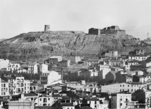 El castell i el turó de Puigterrà, vistos des del sector del Passeig. Aquest sector era conegut popularment com la muntanya del Castell, però en realitat a principi de segle XX solament restaven les ruïnes d'un fortí construït l'any 1822 i ampliat el 1834. Era el fortí de Santa Isabel. Anà caient en desús, fins que amb la tercera carlinada s'utilitzà novament aquesta fortificació. L'any 1880, l'exèrcit l'abandonà definitivament. Destaca la nuesa del turó de Puigterrà, nuesa que en el decurs del primer terç de segle s'intentaria convertir en verdor i, per tant, en parc públic. Però la iniciativa no reeixí. Des d'un punt de vista urbanístic, la continuació de la pujada del Castell i l'alineament del carrer de Circumval·lació daten del darrer decenni del segle passat. Gràcies a aquestes llargues i costoses obres fou possible la comunicació de l'antic camí de Cardona (avui carrer d'Àngel Guimerà) amb la carretera de Vic al voltant del turó. Al parer de Fidel Riu, l'aspecte que oferia aquest sector a l'agost del 1927 era aquest: «Avui, aquelles cases amuntegades sense ordre ni concert, com si s'empaitessin vessant amunt per fugir dels mals tractes manresans, ofereix un aspecte deplorable. Pensem que el turó de Puigterrà dóna davant mateix del Grup Escolar, damunt del Passeig.»
Procedència: Arxiu Jaume Pons i Agulló.