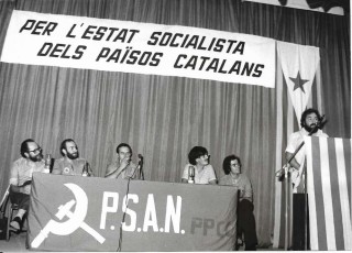 Any 1977. Míting als Carlins. D’esquerra a dreta: Lluís Maria Xirinacs (senador independent), Josep Guia (PSAN), (representant de HASI, partit que va confluir a Herri Batasuna), representant de Su Populu Sardu, Ignasi Perramon (PSAN). Al faristol : Francisco “Ortzi” Letamendia  (Diputat per Euskadiko Ezkerra)